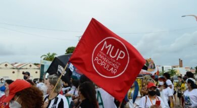 Mulher branca de cabelo loiro, usando blusa do Ceará, segura em meio a outras pessoas, uma bandeira vermelha com um círculo branco e dentro do círculo escrito MUP em letras grandes e logo abaixo Movimento por uma Universidade Popular.