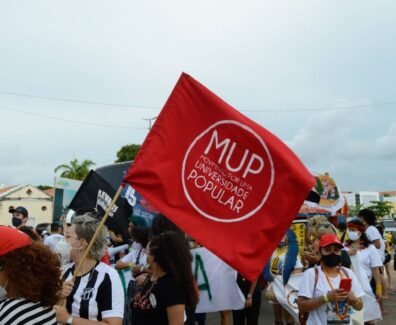 Mulher branca de cabelo loiro, usando blusa do Ceará, segura em meio a outras pessoas, uma bandeira vermelha com um círculo branco e dentro do círculo escrito MUP em letras grandes e logo abaixo Movimento por uma Universidade Popular.