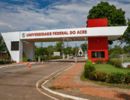 A imagem mostra a entrada da Universidade Federal do Acre, com a inscrição "UNIVERSIDADE FEDERAL DO ACRE" em destaque na parte superior da estrutura branca com detalhes em vermelho. Ao fundo, é possível ver árvores, postes e parte da via interna do campus.