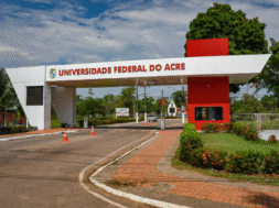 A imagem mostra a entrada da Universidade Federal do Acre, com a inscrição "UNIVERSIDADE FEDERAL DO ACRE" em destaque na parte superior da estrutura branca com detalhes em vermelho. Ao fundo, é possível ver árvores, postes e parte da via interna do campus.