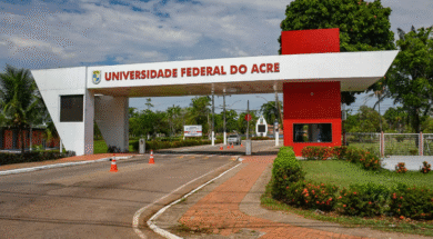 A imagem mostra a entrada da Universidade Federal do Acre, com a inscrição "UNIVERSIDADE FEDERAL DO ACRE" em destaque na parte superior da estrutura branca com detalhes em vermelho. Ao fundo, é possível ver árvores, postes e parte da via interna do campus.