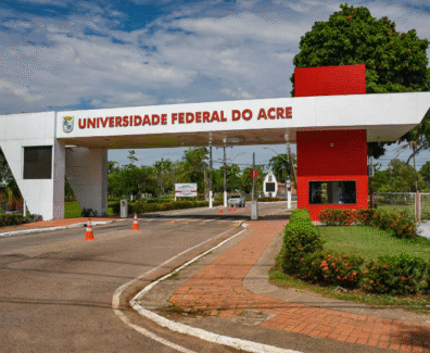 A imagem mostra a entrada da Universidade Federal do Acre, com a inscrição "UNIVERSIDADE FEDERAL DO ACRE" em destaque na parte superior da estrutura branca com detalhes em vermelho. Ao fundo, é possível ver árvores, postes e parte da via interna do campus.