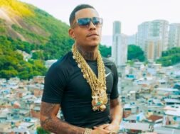 MC Poze posa para câmera com óculos escuros, diversas correntes de ouro no pescoço e uma camisa preta. É um jovem negro com tatuagens no pescoço e bigode. Ao fundo, a favela do Rodo.