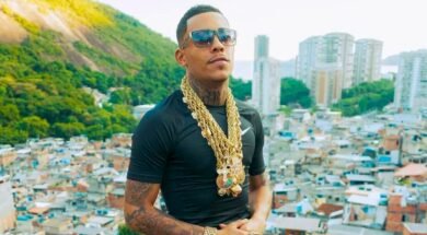 MC Poze posa para câmera com óculos escuros, diversas correntes de ouro no pescoço e uma camisa preta. É um jovem negro com tatuagens no pescoço e bigode. Ao fundo, a favela do Rodo.