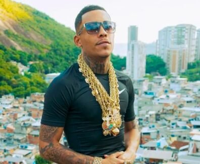 MC Poze posa para câmera com óculos escuros, diversas correntes de ouro no pescoço e uma camisa preta. É um jovem negro com tatuagens no pescoço e bigode. Ao fundo, a favela do Rodo.