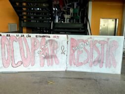 Um quadro branco colocado à frente de uma escada, limitando toda a sua extensão. Nele, está escrito, em vermelho e preto: "Paralização estudantil já! Ocupar e Resistir!". Atrás do quadro, no primeiro lance de escadas, várias cadeiras estão jogadas, sobrepostas umas às outras.