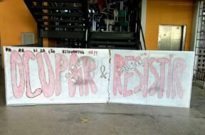 Um quadro branco colocado à frente de uma escada, limitando toda a sua extensão. Nele, está escrito, em vermelho e preto: "Paralização estudantil já! Ocupar e Resistir!". Atrás do quadro, no primeiro lance de escadas, várias cadeiras estão jogadas, sobrepostas umas às outras.