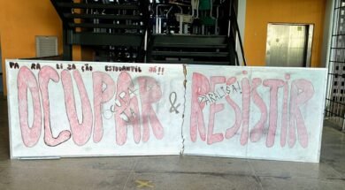 Um quadro branco colocado à frente de uma escada, limitando toda a sua extensão. Nele, está escrito, em vermelho e preto: "Paralização estudantil já! Ocupar e Resistir!". Atrás do quadro, no primeiro lance de escadas, várias cadeiras estão jogadas, sobrepostas umas às outras.