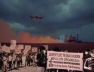 Imagens de manifestações organizadas por trabalhadores sobre a ilustração de uma plantação de milho sendo pulverizada por um drone. À esquerda, uma manifestação contra o agronegócio, com cartazes como: Agrotóxico = morte. À direita, militantes da UJC seguram um cartaz onde está escrito: "Juventude trabalhadora vai à luta organizada. Por nossos direitos, nossos estudos e nossos empregos. Frente de Jovens Trabalhadores.". Mais ao fundo há a lustração de uma usina de enriquecimento de urânio. Créditos: Gabriel Remígio / UJC.