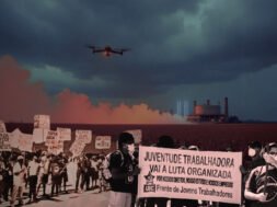 Imagens de manifestações organizadas por trabalhadores sobre a ilustração de uma plantação de milho sendo pulverizada por um drone. À esquerda, uma manifestação contra o agronegócio, com cartazes como: Agrotóxico = morte. À direita, militantes da UJC seguram um cartaz onde está escrito: "Juventude trabalhadora vai à luta organizada. Por nossos direitos, nossos estudos e nossos empregos. Frente de Jovens Trabalhadores.". Mais ao fundo há a lustração de uma usina de enriquecimento de urânio. Créditos: Gabriel Remígio / UJC.