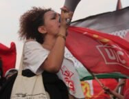 Mulher trans jovem segura um megafone e discursa em uma manifestação, usando camiseta da UJC e bolsa com bandeiras trans e LGBTQIA+, onde se lê “Niterói Capital LGBTQIA+”. Ao fundo, várias bandeiras vermelhas da UJC e do PCB tremulam, compondo um cenário de protesto e resistência popular. Foto: Jonas Amaral (@foto20e1) / UJC.