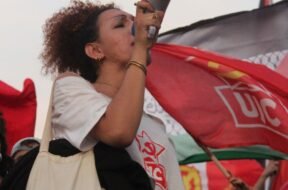 Mulher trans jovem segura um megafone e discursa em uma manifestação, usando camiseta da UJC e bolsa com bandeiras trans e LGBTQIA+, onde se lê “Niterói Capital LGBTQIA+”. Ao fundo, várias bandeiras vermelhas da UJC e do PCB tremulam, compondo um cenário de protesto e resistência popular. Foto: Jonas Amaral (@foto20e1) / UJC.
