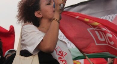 Mulher trans jovem segura um megafone e discursa em uma manifestação, usando camiseta da UJC e bolsa com bandeiras trans e LGBTQIA+, onde se lê “Niterói Capital LGBTQIA+”. Ao fundo, várias bandeiras vermelhas da UJC e do PCB tremulam, compondo um cenário de protesto e resistência popular. Foto: Jonas Amaral (@foto20e1) / UJC.