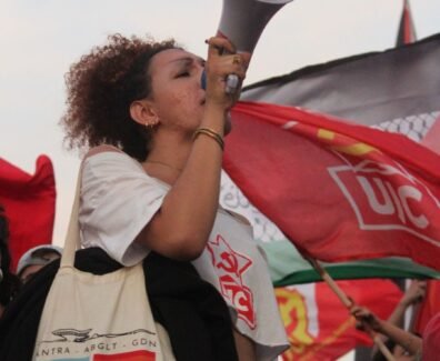 Mulher trans jovem segura um megafone e discursa em uma manifestação, usando camiseta da UJC e bolsa com bandeiras trans e LGBTQIA+, onde se lê “Niterói Capital LGBTQIA+”. Ao fundo, várias bandeiras vermelhas da UJC e do PCB tremulam, compondo um cenário de protesto e resistência popular. Foto: Jonas Amaral (@foto20e1) / UJC.