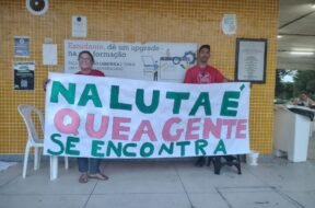 Duas pessoas seguram uma faixa escrita 'Na luta é que a gente se encontra' em frente a uma parede com azulejos amarelos, dentro de uma universidade. Uma delas veste camisa da MUP (Movimento por uma Universidade Popular). Foto: Reprodução / MUP - São Mateus.