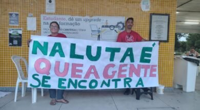 Duas pessoas seguram uma faixa escrita 'Na luta é que a gente se encontra' em frente a uma parede com azulejos amarelos, dentro de uma universidade. Uma delas veste camisa da MUP (Movimento por uma Universidade Popular). Foto: Reprodução / MUP - São Mateus.