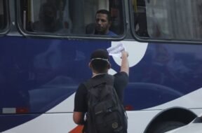 Jovem entregando um panfleto pela janela de um ônibus para um homem.