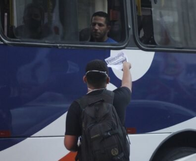 Jovem entregando um panfleto pela janela de um ônibus para um homem.