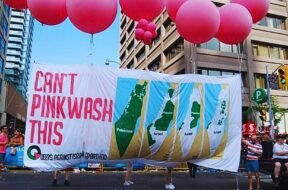 Foto de manifestação com balões rosas e uma grande faixa escrito "Não tem como fazer pinkwash disso", do movimento Queers contra o Apartheid Israelense.