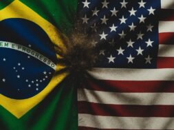 Bandeira brasileira (verde, amarela, azul e branca, com 'Ordem e Progresso') em choque visual contra a bandeira dos EUA (listras vermelhas e brancas, cantão azul com estrelas), gerando um efeito de areia dispersa que simboliza o conflito comercial.