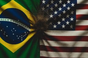 Bandeira brasileira (verde, amarela, azul e branca, com 'Ordem e Progresso') em choque visual contra a bandeira dos EUA (listras vermelhas e brancas, cantão azul com estrelas), gerando um efeito de areia dispersa que simboliza o conflito comercial.