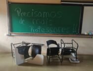 Foto horizontal tirada em uma sala de aula de uma universidade brasileira. No fundo da imagem, há um quadro verde de giz com a frase escrita em letras grandes: “Precisamos de mais professores!!”. A frase ocupa quase todo o quadro, da esquerda para a direita e do topo ao centro. O texto está escrito com giz branco e azul claro. À direita do quadro verde, há parte de um quadro branco fixado na mesma moldura. Sobre o quadro branco, há alguns objetos no canto inferior direito, incluindo uma caixa de lenços de papel e um copo plástico. Em frente ao quadro, no chão, há cinco carteiras escolares viradas de cabeça para baixo. As carteiras têm estrutura metálica preta, encosto e assento pretos com furos circulares, e braço de apoio branco. Algumas estão deitadas sobre o encosto e outras de lado, desalinhadas entre si. O chão é de cor bege brilhante e a parede do fundo é bege, com textura rugosa.