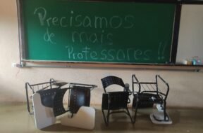 Foto horizontal tirada em uma sala de aula de uma universidade brasileira. No fundo da imagem, há um quadro verde de giz com a frase escrita em letras grandes: “Precisamos de mais professores!!”. A frase ocupa quase todo o quadro, da esquerda para a direita e do topo ao centro. O texto está escrito com giz branco e azul claro. À direita do quadro verde, há parte de um quadro branco fixado na mesma moldura. Sobre o quadro branco, há alguns objetos no canto inferior direito, incluindo uma caixa de lenços de papel e um copo plástico. Em frente ao quadro, no chão, há cinco carteiras escolares viradas de cabeça para baixo. As carteiras têm estrutura metálica preta, encosto e assento pretos com furos circulares, e braço de apoio branco. Algumas estão deitadas sobre o encosto e outras de lado, desalinhadas entre si. O chão é de cor bege brilhante e a parede do fundo é bege, com textura rugosa.