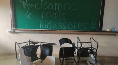 Foto horizontal tirada em uma sala de aula de uma universidade brasileira. No fundo da imagem, há um quadro verde de giz com a frase escrita em letras grandes: “Precisamos de mais professores!!”. A frase ocupa quase todo o quadro, da esquerda para a direita e do topo ao centro. O texto está escrito com giz branco e azul claro. À direita do quadro verde, há parte de um quadro branco fixado na mesma moldura. Sobre o quadro branco, há alguns objetos no canto inferior direito, incluindo uma caixa de lenços de papel e um copo plástico. Em frente ao quadro, no chão, há cinco carteiras escolares viradas de cabeça para baixo. As carteiras têm estrutura metálica preta, encosto e assento pretos com furos circulares, e braço de apoio branco. Algumas estão deitadas sobre o encosto e outras de lado, desalinhadas entre si. O chão é de cor bege brilhante e a parede do fundo é bege, com textura rugosa.