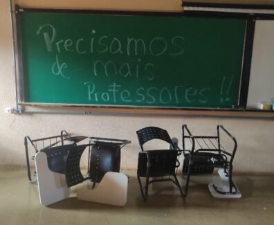 Foto horizontal tirada em uma sala de aula de uma universidade brasileira. No fundo da imagem, há um quadro verde de giz com a frase escrita em letras grandes: “Precisamos de mais professores!!”. A frase ocupa quase todo o quadro, da esquerda para a direita e do topo ao centro. O texto está escrito com giz branco e azul claro. À direita do quadro verde, há parte de um quadro branco fixado na mesma moldura. Sobre o quadro branco, há alguns objetos no canto inferior direito, incluindo uma caixa de lenços de papel e um copo plástico. Em frente ao quadro, no chão, há cinco carteiras escolares viradas de cabeça para baixo. As carteiras têm estrutura metálica preta, encosto e assento pretos com furos circulares, e braço de apoio branco. Algumas estão deitadas sobre o encosto e outras de lado, desalinhadas entre si. O chão é de cor bege brilhante e a parede do fundo é bege, com textura rugosa.