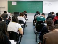 Fotografia retangular horizontal de uma sala de aula com aproximadamente vinte estudantes sentados em cadeiras com pranchetas, de costas para a câmera, fazendo uma prova. A maioria está inclinada para frente escrevendo ou lendo. As cadeiras são pretas, e o piso é cinza. Ao fundo, há um quadro verde com escritos em giz branco e colorido, com os dizeres, centralizados: "Duração total da prova: 5h. Início: 13h22min. Término: 18h22. Saída permitida a partir das 15h22". Acima do quadro, à esquerda, há um cartaz branco com moldura vermelha e texto em vermelho e azul, onde se lê: "A devolução do cartão-resposta é obrigatória". À direita do quadro verde há um quadro branco com dois avisos iguais, lado a lado, com texto em letras pretas, listando itens proibidos no dia da prova. Algumas garrafas de água estão no chão, ao lado de carteiras. Foto: Marcos Santos / USP Imagens.