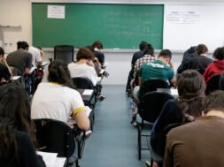Fotografia retangular horizontal de uma sala de aula com aproximadamente vinte estudantes sentados em cadeiras com pranchetas, de costas para a câmera, fazendo uma prova. A maioria está inclinada para frente escrevendo ou lendo. As cadeiras são pretas, e o piso é cinza. Ao fundo, há um quadro verde com escritos em giz branco e colorido, com os dizeres, centralizados: "Duração total da prova: 5h. Início: 13h22min. Término: 18h22. Saída permitida a partir das 15h22". Acima do quadro, à esquerda, há um cartaz branco com moldura vermelha e texto em vermelho e azul, onde se lê: "A devolução do cartão-resposta é obrigatória". À direita do quadro verde há um quadro branco com dois avisos iguais, lado a lado, com texto em letras pretas, listando itens proibidos no dia da prova. Algumas garrafas de água estão no chão, ao lado de carteiras. Foto: Marcos Santos / USP Imagens.