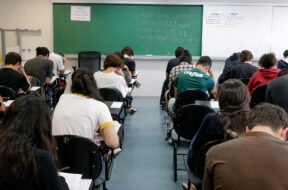 Fotografia retangular horizontal de uma sala de aula com aproximadamente vinte estudantes sentados em cadeiras com pranchetas, de costas para a câmera, fazendo uma prova. A maioria está inclinada para frente escrevendo ou lendo. As cadeiras são pretas, e o piso é cinza. Ao fundo, há um quadro verde com escritos em giz branco e colorido, com os dizeres, centralizados: "Duração total da prova: 5h. Início: 13h22min. Término: 18h22. Saída permitida a partir das 15h22". Acima do quadro, à esquerda, há um cartaz branco com moldura vermelha e texto em vermelho e azul, onde se lê: "A devolução do cartão-resposta é obrigatória". À direita do quadro verde há um quadro branco com dois avisos iguais, lado a lado, com texto em letras pretas, listando itens proibidos no dia da prova. Algumas garrafas de água estão no chão, ao lado de carteiras. Foto: Marcos Santos / USP Imagens.