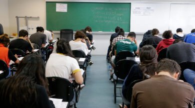 Fotografia retangular horizontal de uma sala de aula com aproximadamente vinte estudantes sentados em cadeiras com pranchetas, de costas para a câmera, fazendo uma prova. A maioria está inclinada para frente escrevendo ou lendo. As cadeiras são pretas, e o piso é cinza. Ao fundo, há um quadro verde com escritos em giz branco e colorido, com os dizeres, centralizados: "Duração total da prova: 5h. Início: 13h22min. Término: 18h22. Saída permitida a partir das 15h22". Acima do quadro, à esquerda, há um cartaz branco com moldura vermelha e texto em vermelho e azul, onde se lê: "A devolução do cartão-resposta é obrigatória". À direita do quadro verde há um quadro branco com dois avisos iguais, lado a lado, com texto em letras pretas, listando itens proibidos no dia da prova. Algumas garrafas de água estão no chão, ao lado de carteiras. Foto: Marcos Santos / USP Imagens.