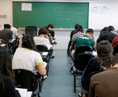 Fotografia retangular horizontal de uma sala de aula com aproximadamente vinte estudantes sentados em cadeiras com pranchetas, de costas para a câmera, fazendo uma prova. A maioria está inclinada para frente escrevendo ou lendo. As cadeiras são pretas, e o piso é cinza. Ao fundo, há um quadro verde com escritos em giz branco e colorido, com os dizeres, centralizados: "Duração total da prova: 5h. Início: 13h22min. Término: 18h22. Saída permitida a partir das 15h22". Acima do quadro, à esquerda, há um cartaz branco com moldura vermelha e texto em vermelho e azul, onde se lê: "A devolução do cartão-resposta é obrigatória". À direita do quadro verde há um quadro branco com dois avisos iguais, lado a lado, com texto em letras pretas, listando itens proibidos no dia da prova. Algumas garrafas de água estão no chão, ao lado de carteiras. Foto: Marcos Santos / USP Imagens.