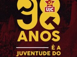 Imagem quadrada com fundo em tons escuros de vermelho e preto com textura pontilhada. Ao centro da imagem, em letras grandes amarelas, está escrito "98". O número "9" está à esquerda e o "8" à direita. Dentro do número "8", no topo, há o símbolo da UJC: uma estrela vermelha com foice e martelo amarelos, e abaixo dela um retângulo vermelho com as letras brancas "UJC". Abaixo dos números, centralizado, está escrito em letras amarelas: "ANOS". Mais abaixo, também centralizado, aparece a frase "É A JUVENTUDE DO PCB", em que "É A" e "JUVENTUDE DO" estão em letras amarelas e "PCB" está em letras amarelas acompanhadas do símbolo da foice e martelo à esquerda da sigla. Acima e abaixo da frase há linhas horizontais brancas.