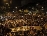 Fotografia de uma manifestação noturna em uma avenida. A imagem mostra uma multidão de pessoas ocupando toda a rua, segurando cartazes e faixas. Em primeiro plano, uma faixa branca com letras pretas está esticada horizontalmente e diz: "NÃO SE INTIMIDAR, NÃO DESMOBILIZAR! RODEAR DE SOLIDARIEDADE OS QUE LUTAM!". Outras faixas e cartazes aparecem mais ao fundo, mas não estão legíveis com clareza. As pessoas ocupam toda a largura da via, e ao fundo há postes de iluminação pública, árvores e prédios.