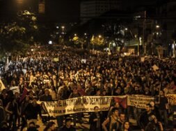 Fotografia de uma manifestação noturna em uma avenida. A imagem mostra uma multidão de pessoas ocupando toda a rua, segurando cartazes e faixas. Em primeiro plano, uma faixa branca com letras pretas está esticada horizontalmente e diz: "NÃO SE INTIMIDAR, NÃO DESMOBILIZAR! RODEAR DE SOLIDARIEDADE OS QUE LUTAM!". Outras faixas e cartazes aparecem mais ao fundo, mas não estão legíveis com clareza. As pessoas ocupam toda a largura da via, e ao fundo há postes de iluminação pública, árvores e prédios.