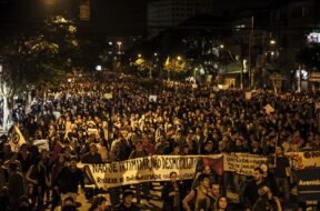 Fotografia de uma manifestação noturna em uma avenida. A imagem mostra uma multidão de pessoas ocupando toda a rua, segurando cartazes e faixas. Em primeiro plano, uma faixa branca com letras pretas está esticada horizontalmente e diz: "NÃO SE INTIMIDAR, NÃO DESMOBILIZAR! RODEAR DE SOLIDARIEDADE OS QUE LUTAM!". Outras faixas e cartazes aparecem mais ao fundo, mas não estão legíveis com clareza. As pessoas ocupam toda a largura da via, e ao fundo há postes de iluminação pública, árvores e prédios.
