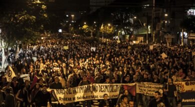 Fotografia de uma manifestação noturna em uma avenida. A imagem mostra uma multidão de pessoas ocupando toda a rua, segurando cartazes e faixas. Em primeiro plano, uma faixa branca com letras pretas está esticada horizontalmente e diz: "NÃO SE INTIMIDAR, NÃO DESMOBILIZAR! RODEAR DE SOLIDARIEDADE OS QUE LUTAM!". Outras faixas e cartazes aparecem mais ao fundo, mas não estão legíveis com clareza. As pessoas ocupam toda a largura da via, e ao fundo há postes de iluminação pública, árvores e prédios.