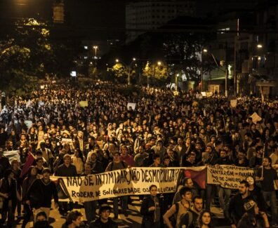 Fotografia de uma manifestação noturna em uma avenida. A imagem mostra uma multidão de pessoas ocupando toda a rua, segurando cartazes e faixas. Em primeiro plano, uma faixa branca com letras pretas está esticada horizontalmente e diz: "NÃO SE INTIMIDAR, NÃO DESMOBILIZAR! RODEAR DE SOLIDARIEDADE OS QUE LUTAM!". Outras faixas e cartazes aparecem mais ao fundo, mas não estão legíveis com clareza. As pessoas ocupam toda a largura da via, e ao fundo há postes de iluminação pública, árvores e prédios.