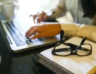 Fotografia em ambiente interno. Em primeiro plano, sobre uma mesa preta, há um caderno de capa branca com espiral preto. Sobre o caderno, um par de óculos de grau com armação preta está apoiado. Ao fundo, à esquerda, aparece parcialmente um notebook prateado com teclado preto, sendo utilizado por uma pessoa. Apenas as mãos e parte do braço direito dessa pessoa estão visíveis, digitando no teclado. À direita, desfocada, vê-se parte do cabelo e da blusa clara da pessoa. No canto superior esquerdo, há uma xícara branca sobre a mesa. Foto: Marcelo Camargo / Agência Brasil.