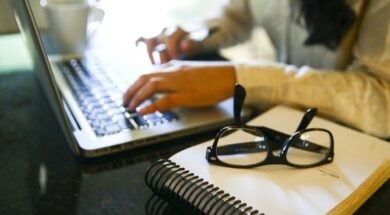 Fotografia em ambiente interno. Em primeiro plano, sobre uma mesa preta, há um caderno de capa branca com espiral preto. Sobre o caderno, um par de óculos de grau com armação preta está apoiado. Ao fundo, à esquerda, aparece parcialmente um notebook prateado com teclado preto, sendo utilizado por uma pessoa. Apenas as mãos e parte do braço direito dessa pessoa estão visíveis, digitando no teclado. À direita, desfocada, vê-se parte do cabelo e da blusa clara da pessoa. No canto superior esquerdo, há uma xícara branca sobre a mesa. Foto: Marcelo Camargo / Agência Brasil.