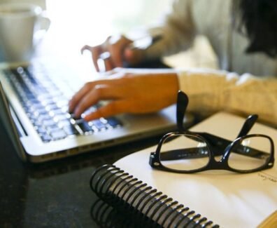 Fotografia em ambiente interno. Em primeiro plano, sobre uma mesa preta, há um caderno de capa branca com espiral preto. Sobre o caderno, um par de óculos de grau com armação preta está apoiado. Ao fundo, à esquerda, aparece parcialmente um notebook prateado com teclado preto, sendo utilizado por uma pessoa. Apenas as mãos e parte do braço direito dessa pessoa estão visíveis, digitando no teclado. À direita, desfocada, vê-se parte do cabelo e da blusa clara da pessoa. No canto superior esquerdo, há uma xícara branca sobre a mesa. Foto: Marcelo Camargo / Agência Brasil.