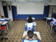 Sala de aula com piso de ladrilhos bege estampados em marrom. No fundo da sala há uma lousa branca grande com alguns escritos, cercada por paredes pintadas de azul na parte inferior e branca na parte superior. Acima da lousa, está presa uma faixa com desenhos coloridos. À esquerda, há cartazes coloridos colados na parede, e à direita, dois painéis de papel pardo com atividades. Sete crianças estão sentadas individualmente em carteiras azuis, todas de costas para a câmera. Elas usam uniforme azul claro com detalhes em azul escuro e estão escrevendo em cadernos sobre as mesas. Foto: Tânia Rêgo / Agência Brasil.