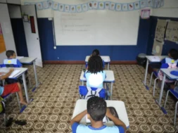 Sala de aula com piso de ladrilhos bege estampados em marrom. No fundo da sala há uma lousa branca grande com alguns escritos, cercada por paredes pintadas de azul na parte inferior e branca na parte superior. Acima da lousa, está presa uma faixa com desenhos coloridos. À esquerda, há cartazes coloridos colados na parede, e à direita, dois painéis de papel pardo com atividades. Sete crianças estão sentadas individualmente em carteiras azuis, todas de costas para a câmera. Elas usam uniforme azul claro com detalhes em azul escuro e estão escrevendo em cadernos sobre as mesas. Foto: Tânia Rêgo / Agência Brasil.