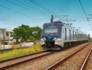 A imagem mostra o metrô em uma das ferrovias do Recife, com casas ao lado esquerdo. Na parte inferior, há gramados e capins em tons de verde e amarelo. Acima, um céu azul indica que possivelmente já seja o período da tarde.