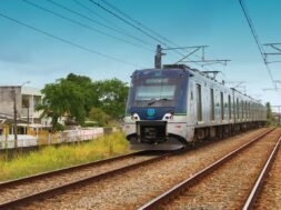 A imagem mostra o metrô em uma das ferrovias do Recife, com casas ao lado esquerdo. Na parte inferior, há gramados e capins em tons de verde e amarelo. Acima, um céu azul indica que possivelmente já seja o período da tarde.
