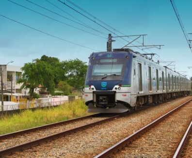 A imagem mostra o metrô em uma das ferrovias do Recife, com casas ao lado esquerdo. Na parte inferior, há gramados e capins em tons de verde e amarelo. Acima, um céu azul indica que possivelmente já seja o período da tarde.