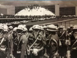 Fotografia em preto e branco. Em primeiro plano, um grupo de militares ocupa a parte inferior da imagem. Eles vestem uniformes escuros, capacetes brancos, cintos e bandoleiras brancas. Usam também máscaras ou lenços cobrindo o rosto. Ao fundo, há um grande número de pessoas reunidas em frente e sobre a cúpula branca do Congresso Nacional, em Brasília. Atrás da cúpula, aparecem os prédios retangulares das duas torres do Congresso. Diversos automóveis antigos estão estacionados ao longo da via em frente ao prédio. O céu está parcialmente nublado.