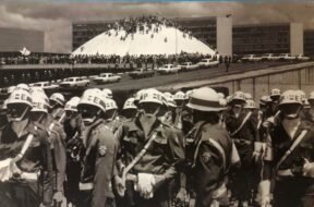 Fotografia em preto e branco. Em primeiro plano, um grupo de militares ocupa a parte inferior da imagem. Eles vestem uniformes escuros, capacetes brancos, cintos e bandoleiras brancas. Usam também máscaras ou lenços cobrindo o rosto. Ao fundo, há um grande número de pessoas reunidas em frente e sobre a cúpula branca do Congresso Nacional, em Brasília. Atrás da cúpula, aparecem os prédios retangulares das duas torres do Congresso. Diversos automóveis antigos estão estacionados ao longo da via em frente ao prédio. O céu está parcialmente nublado.